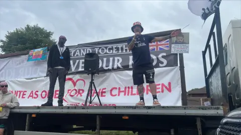 Hundreds of protesters gather at asylum hotel in Norwich area Richard Knights/BBC A man in black T-shirt and shorts and wearing a union jack bucket hat addresses a crowd from the back of a lorry. Another man stands beside him and looks on. Behind them are banners, one of which says "Unite as one for our children".