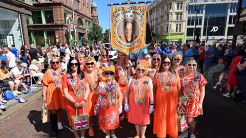 Thousands attending NI's Orange Order parades Pacemaker A group of women in orange dresses and sashes wearing sunglasses as they pose on a street in Belfast and hundreds of spectators are behind them. The women are holding a banner that says "Sister Clarke Memorial L.L.O.L No. 156 Parkhead Dist 2". The sun is shining on the women.