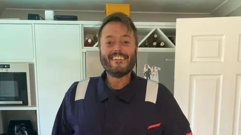 Military aircrew pursuing MoD after cancer diagnoses reaches 180 Anna-Louise Stubbings Zach standing in a kitchen wearing a blue military boiler suit and smiling at the camera. He has a short beard and brown hair.