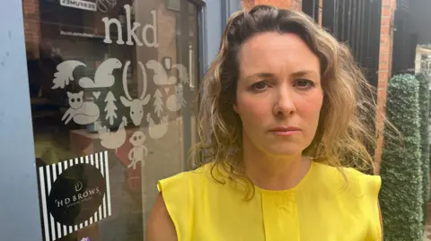Salon owner 'ready' for key L'Oréal trademark dispute hearing A woman wearing a bright yellow top standing in front of a door with nkd branding on it.