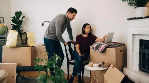Lifetime ISAs: Why they divide opinion Getty Images Man and woman look at laptop in a living room surrounded by cardboard boxes. The woman is sitting on a dining chair, the man is standing behind her, leaning on the back of her chair, the laptop is on top of a big cardboard box.