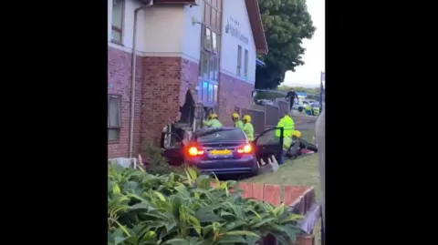 Arrests as Sunderland care home crash puts eight in hospital SGM A blue BMW car with its doors open and its rear headlights on, halfway through a brick building. Firemen in hi-vis jackets surround the car.