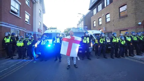 Epping council votes to urge government to shut asylum hotel PA Media A man holds a St George's flag standing in front of a line of armed police officers with yellow high-vis jackets and face shields. There are flats either side of them and police vans behind them.