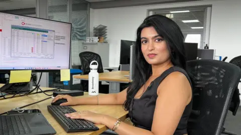 Defence firms 'confident' hundreds more jobs will come A woman in a black top, with black hair, sits at a desk. She is looking into the camera. Her hands are on a keyboard and mouse, and a computer monitor is behind her.