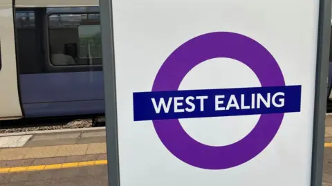 Ealing tenants experience steepest rise in London rent A white sign with a grey rim on the right displays Transport for London's roundel, branded in the colours of the Elizabeth line. There is a hollow purple circle, with a dark blue box running horizontally across the middle containing writing reading 'West Ealing'. Behind the sign, a train platform is visible in the background. There is a yellow line running along the platform. A train is stationed at the platform, coloured in purple, black and white.
