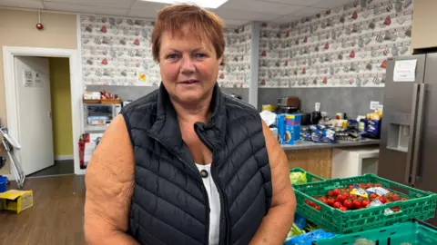 'Food demand in Cumbria is unprecedented' Diane Snedker, wearing a black gilet, with short red hair, stands next to boxes of food in a kitchen.
