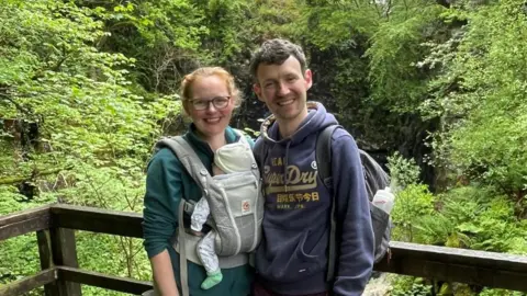 Lifetime ISAs: Why they divide opinion Lucy Slavin Lucy and Daniel Slavin stand with woodland seen behind them. Lucy is carrying their young baby in a baby carrier on her chest.