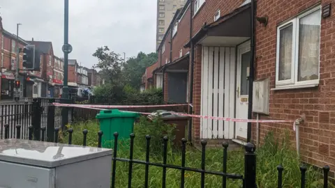 How did no-one know about mum and daughter's death for months? The front of the house with red and white tape around it, overgrown grass, a green and brown bin