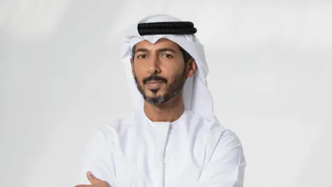 The Gulf bets big on AI as it seeks the 'new oil' Khazna Hassan Alnaqbi Hassan looks at the camera with his arms crossed. He is wearing the traditional emirati dress - a Kandura - a long white robe.
On his head he wears a white headdress called ghutra and secured in place by a black cord called the agal.