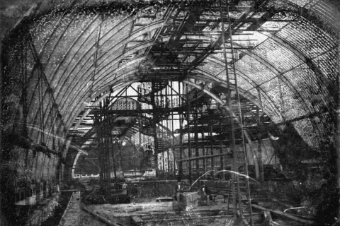 Palm House set for major renovation RBG Kew A black and white image taken inside the Palm House, a glass and iron building in Kew Gardens. It shows it part way though construction. Scaffolding and tall ladders run from the floor to the ceiling.