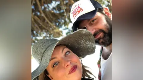 'Fantasist' promised music stars for festival that never happened Kate Kate, on the left, is smiling into the camera. She has blue eyes, dark blonde hair and pink lipstick. She is wearing a floppy straw hat that is partially covering the face of her partner James, who has a dark beard, white T-shirt and a cap with the words 'everybody gotta thing’.