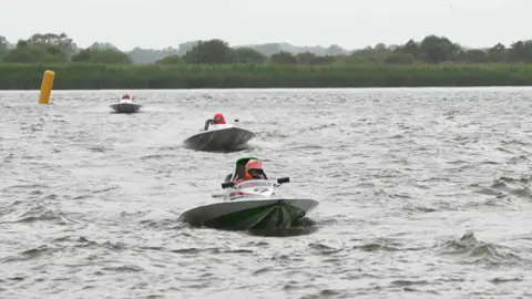 Powerboat driver dies after crash with houseboat at Oulton Broad BBC Three powerboats whizz towards the camera on a choppy river, with marsh and trees in the background. The three drivers wear orange helmets.