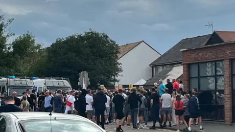 Officers attacked at Epping asylum hotel protest, police say BBC A large group of people walking and gathering outside buildings. Most are wearing black and one person has a St George's flag draped over their shoulders. There are two police vans visible behind the group in the background.