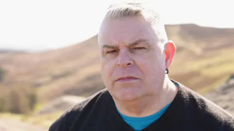 Police investigate sex abuse claims against men linked to Church Alisdair does not smile as he stands near some rocks with a hill behind him. He is wearing a bright blue t-shirt under a dark v-neck jumper and has an earing in his left ear. His short white hair is brushed back.