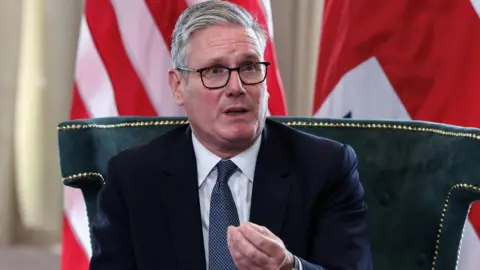 Reuters Sir Keir Starmer, sits on a green upholstered chair with gold trim, wearing a dark suit, white shirt, and blue patterned tie. He gestures with his right hand.