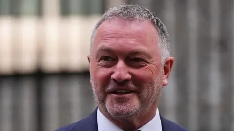 Water company sewage pollution to halve by 2030, minister pledges PA Media Environment Secretary Steve Reed. He is smiling and wearing a navy blue suit jacket with a white collared shirt