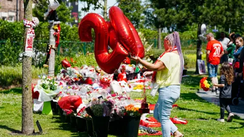 PA Media A woman in a yellow top and jeans, with her hair in long purple braids, places ballons in the shape of the number 20 on a grassy area blanketed in flowers, football shirts, scarves and balloons