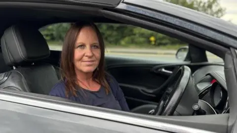 Airport drivers try to beat drop-off charges by parking in laybys BBC Caroline O'Brien sitting in her car looking directly at the camera. She is wearing a blue top. She is visible from the shoulders up and is sitting in the driver's seat with the steering wheel in front of her. There is a road and foliage in the background.