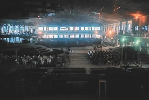 Inside Synagogue Church of all Nations in Lagos, founded by TB Joshua. It is a vast hall with a raised platform and rows of chairs. 
