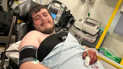 Cardiff man says triple dog attack has left him with nightmares William Newbury William, with dark hair and a beard, lies in an ambulance. He is covered by a blanket and has wires from machines connected to his body. His hand is covered in blood. He is smiling towards the camera.