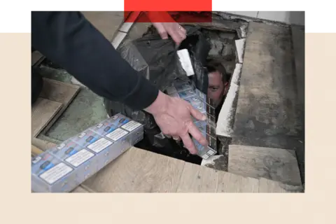 The illegal cigarettes trade in the UK signals a deeper problem BBC/Phillip Edwards An officer bring out illegal packets of cigarettes from a hole in the floor following a raid on a corner shop