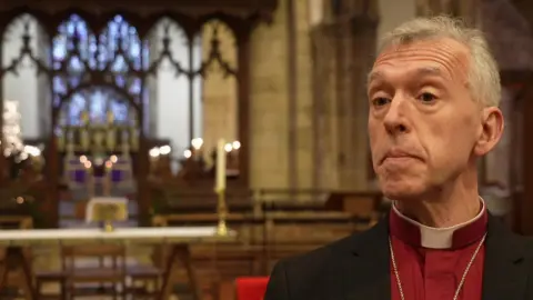 Bangor Cathedral had 'binge drinking culture' at diocese A picture of the outgoing Archbishop of Wales Andrew John wearing a purple top and a dog collar with a chain around his neck inside a cathedrawl with candles lit in the background