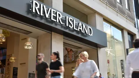 Getty Images Shoppers walk past a River Island store on Oxford Street in London. It's a sunny day. Two friends are walking side by side and a woman with short blonde hair is walking.