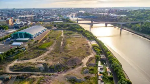 Hi-Track Aerial Photography An aerial view of Forth Yard in Newcastle. The land stands between the River Tyne and the green Utilita Arena. The land is covered in sparse grass and dirt tracks.