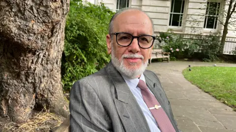 Olympics joy turned into a barbaric loss of life BBC George Psaradakis in a mauve tie, grey suit and black glasses sits on a bench in a park with a tree trunk behind him.