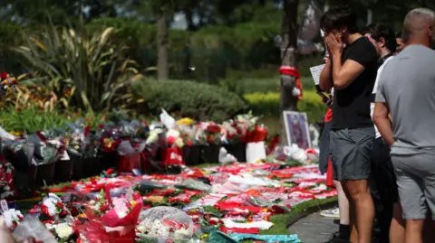 EPA A crowd of people, including a man with his face in his hands, stand on the edge of a grassy area blanketed in flowers, football shirts, scarves and balloons
