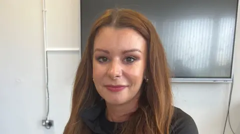 BBC  A head and shoulders shot of Seana McConville who has long brown hair. She is wearing a dark top and has pink lipstick.