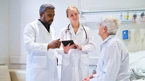 Physician associates need new job title, says review Getty Images Two healthcare professionals wearing white coats, with one holding a tablet, talk to a patient with grey hair who is sitting on a hospital bed
