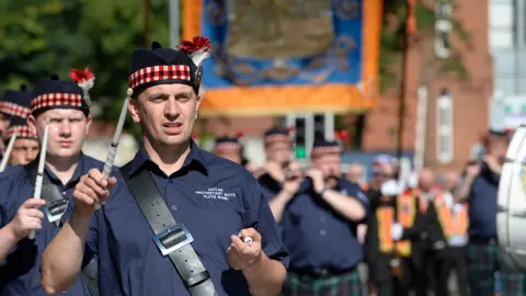 Thousands attending NI's Orange Order parades Pacemaker Members of a band parading down the street wearing navy short sleeved shirts and tartan bordered hats with a red flower. The sun is shining on the band members.