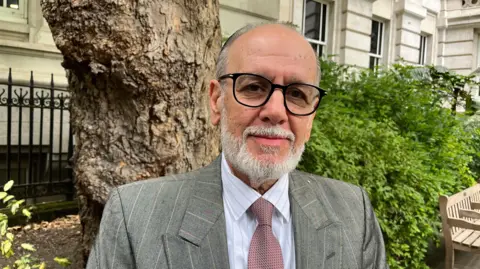 Olympics joy turned into a barbaric loss of life George Psaradakis in a mauve tie, grey suit and black glasses standing in a park with a tree trunk behind him.