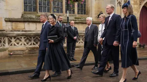 Funeral held for Norman Tebbit at Bury St Edmunds cathedral PA Media Lord Tebbit's three children walking down a street outside the cathedral holding hands with their partners. Behind them are two members of staff.