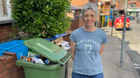 No end in sight for Birmingham bin strike after six months A woman with short grey hair wearing a blue T-shirt that says 'binfluencer' on it. She is stood next to an overflowing green recycling bin on a residential street.
