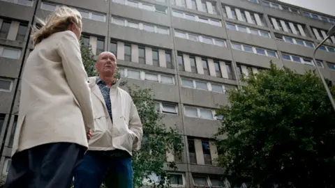 Trainspotting author Irvine Welsh says AI has made us dumbed down machines Adam Walker/BBC Katie Razzall with blond hair, white jacket and trousers talking to Irvine Welsh, bald, in white anorak and jeans outside the Banana flats in Leith