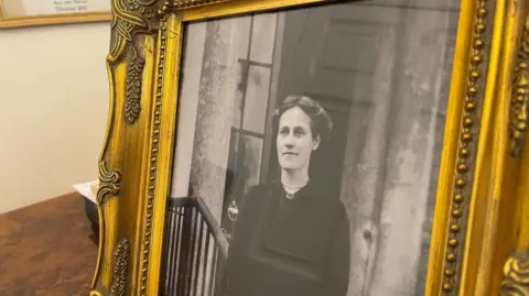 230 years of unbroken weather recordings A framed black and white photo of Theresa Hardcastle. She has long hair which has been parted and tied up around her head. She is wearing a black dress and is standing on the steps of a large building, leaning one arm on a bannister.