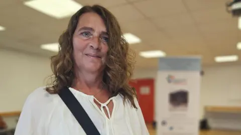 Aisha Iqbal/BBC A middle-aged woman with dark blonde hair and blue eyes stands in what looks like a community hall. She is wearing a white cotton shirt.