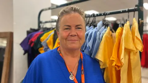 Hard-hit families hail £1 school uniform sale in Wigan Jasmine DuFraisse/BBC Louise Atherton has brown greying hair and is in a blue top in front of a rail school uniform shirts