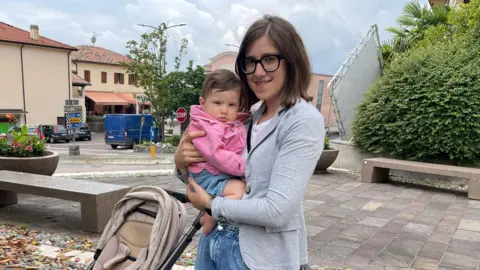 'The village will die' - Italy looks for answers to decline in number of babies Valentina, a woman with dark hair and glasses, holds a baby in a pink jumper.