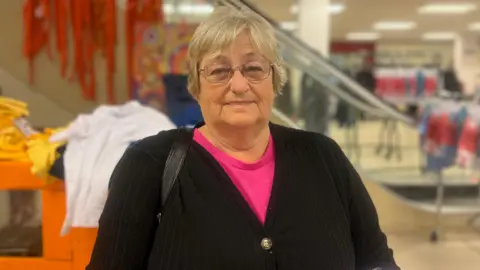 Hard-hit families hail £1 school uniform sale in Wigan Jasmine DuFraisse/BBC Sandra Turton is wearing glasses, has short grey hair and is wearing a pink top with a black cardigan.