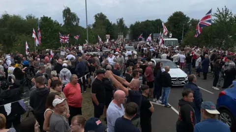Hundreds of protesters gather at asylum hotel in Norwich area Alex Dunlop/BBC A large crowd of people, many waving flags and standing in the road.