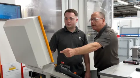 Defence firms 'confident' hundreds more jobs will come Two men in t shirts look at a screen in a factory. One is older. The older man is pointing at the screen.