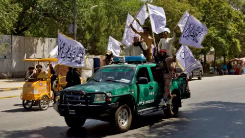 Russia the first to recognise Taliban government in Afghanistan Reuters A battered green police vehicle is driving down a tree-lined street, with five men hanging off the sides waving white flags with Arabic text on them. The men are cheering and wearing elements of military dress, and two of them have large guns strapped to their chests.