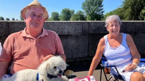 Thousands attending NI's Orange Order parades Terence and Mildred sitting in the sun on the street with their white dog Alfie. Terence is shading his head with a straw hat.