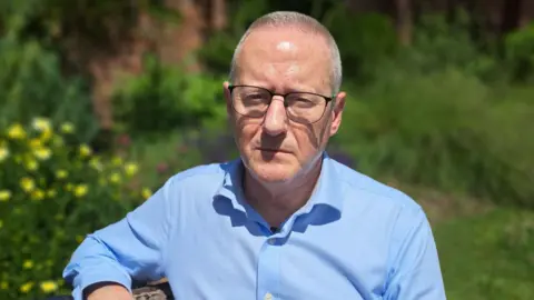 Military aircrew pursuing MoD after cancer diagnoses reaches 180 Graham Jones is sitting on a park bench in a park, flowers can be seen behind him. He has short grey hair, glasses and wears a blue shirt which is open at the collar.