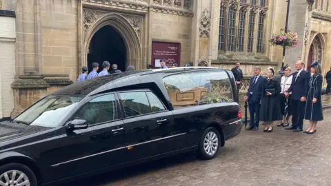 Funeral held for Norman Tebbit at Bury St Edmunds cathedral Luke Deal/BBC A black hearse is parked outside St Edmundsbury Cathedral. A coffin is visible inside with flowers on it. Behind the hearse are family members of Lord Tebbit.
