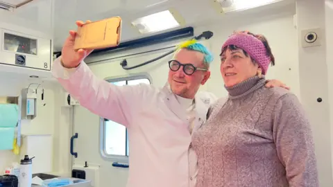 The doctor fighting for women's health on Ukraine's front line Serhii Baksheiev A doctor smiles as he takes a selfie with a woman patient inside a mobile medical unit. His hair is dyed the blue and yellow of the Ukrainian flag, and medical paraphernalia is in the background.