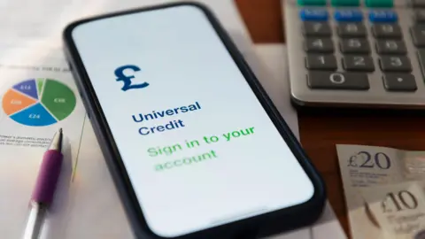 Getty Images A shot of several objects including a pen, a smartphone open on a page to log in to a universal credit account, a calculator and two cash notes of £20 and £10 respectively. 